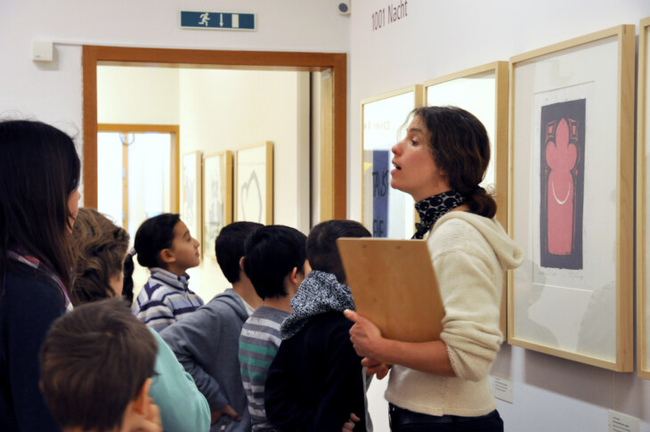Lehrperson steht vor Kunstwerk und erklärt einer Gruppe von Kindern während einer Erlebnisführung etwas.