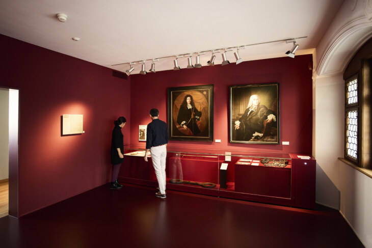 Blick in den Raum Grosser und Kleiner Rat. Der Raum ist rot gestrichen und an der Wand hängen zwei grosse Ölportraits zweier Männer.