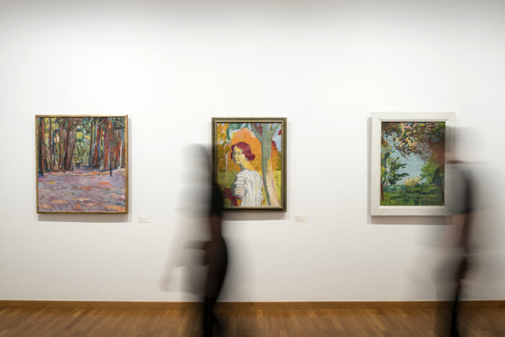 An einer Wand hängen drei sehr farbenfrohe Gemälde von Cuno Amiet. Davor laufen zwei Personen nach links. Es ist aber nur ein schwarzer Schatten zu sehen.