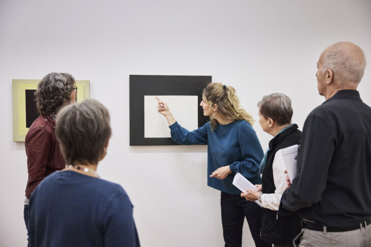 Eine Gruppe steht vor einem abstrakten Kunstgemälde. Eine Frau verweist auf etwas auf dem Bild.