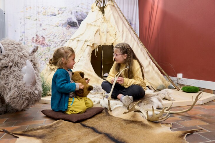 Zwei Kinder sitzen im Steinzeitlager im Museum vor einem Steinzeitzelt.